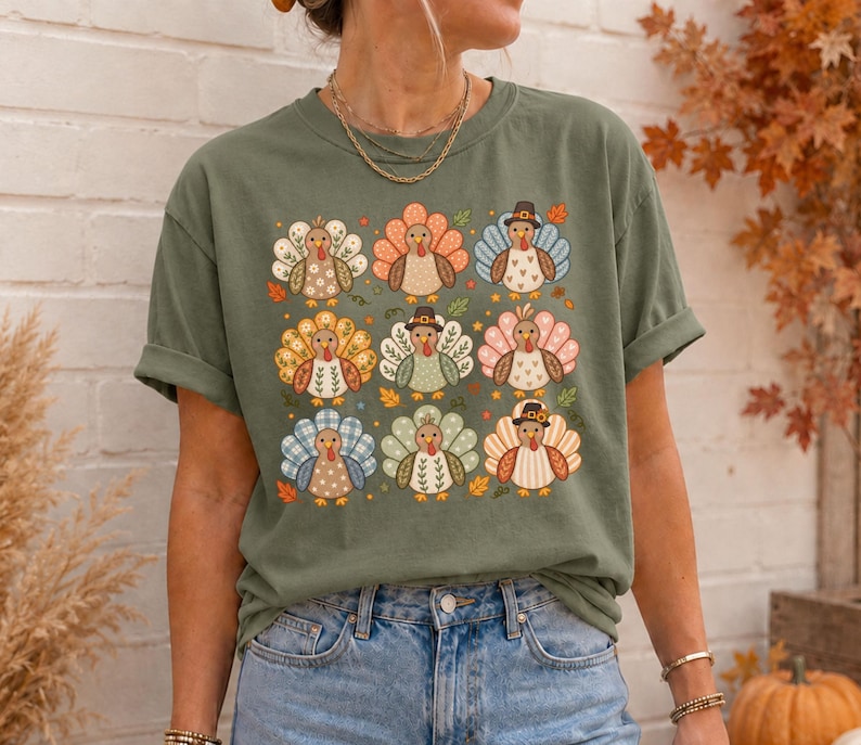 May include: An olive green t-shirt featuring a grid of nine stylized turkey illustrations. Each turkey has a unique patterned tail, including floral, plaid, and polka dot designs, with some wearing small hats, surrounded by scattered autumn leaves.