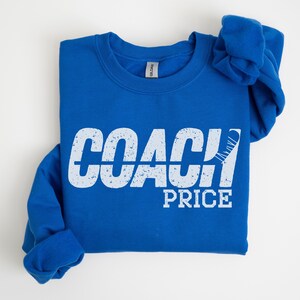 May include: A royal blue sweatshirt with the text "COACH PRICE" in white with a distressed look. A white hockey stick is partially visible behind the word "COACH".