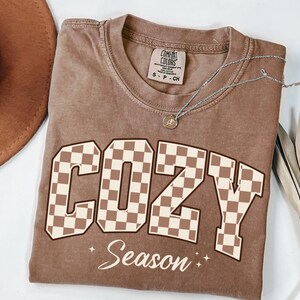 May include: A brown short-sleeved t-shirt with a crew neckline featuring the word COZY in large, brown and cream chequered block letters, with the word Season written in cursive below it, accompanied by two small four-pointed star icons.
