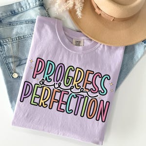 May include: A light purple t-shirt with the words "PROGRESS PERFECTION" in colorful, bubble letters. The shirt is folded on top of a pair of light blue jeans, with a gold necklace and a tan hat in the background.