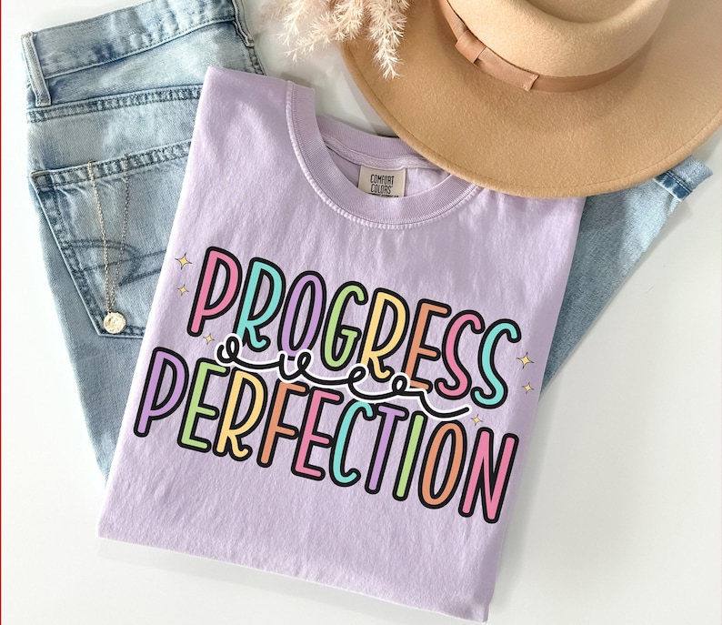 May include: A light purple t-shirt with the words "PROGRESS PERFECTION" in colorful, bubble letters. The shirt is folded on top of a pair of light blue jeans, with a gold necklace and a tan hat in the background.