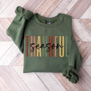 Puede incluir: Una sudadera verde con el texto "Thankful Season" en rayas verticales de colores. El texto est&aacute; en negro.