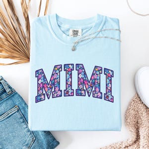 Puede incluir: Una camiseta Comfort Colors azul claro con la palabra "MIMI" en un dise&ntilde;o floral. Las letras est&aacute;n rellenas con un colorido patr&oacute;n de flores en tonos rosa, morado y azul. Un collar plateado con un peque&ntilde;o colgante est&aacute; sobre la camiseta.