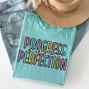 May include: A light blue t-shirt with the words "PROGRESS PERFECTION" in colorful letters. The shirt is folded on top of a pair of light blue jeans, with a tan hat and a gold necklace visible in the background.