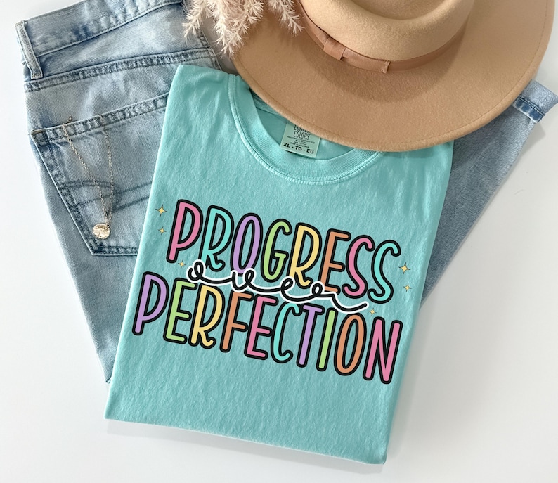 May include: A light blue t-shirt with the words "PROGRESS PERFECTION" in colorful letters. The shirt is folded on top of a pair of light blue jeans, with a tan hat and a gold necklace visible in the background.