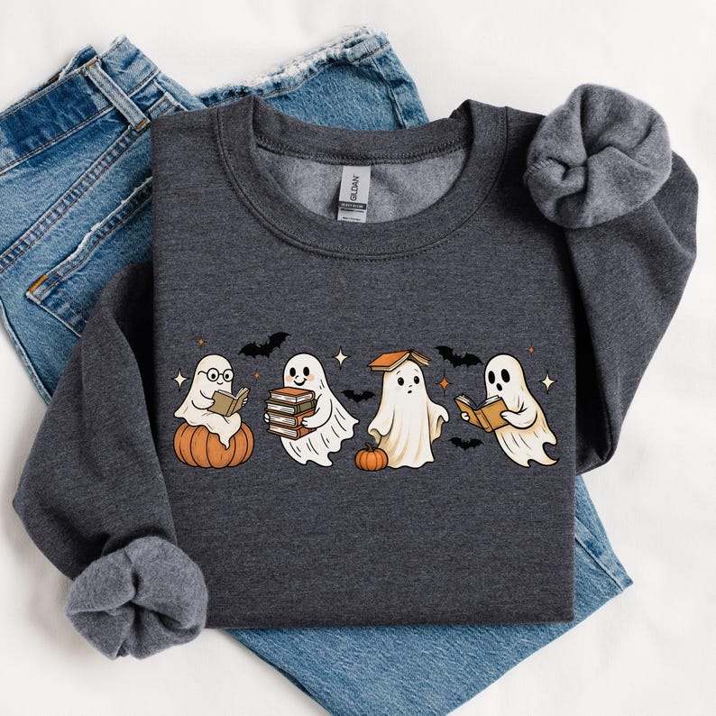 Puede incluir: Sudadera gris oscuro con un dise&ntilde;o de Halloween. El dise&ntilde;o incluye cuatro fantasmas de dibujos animados leyendo libros, con calabazas, murci&eacute;lagos y estrellas. La sudadera est&aacute; doblada sobre unos vaqueros azules.