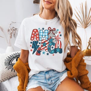 May include: White t-shirt featuring a colourful "AMERICA" design. The graphic includes stars, stripes, and a lightning bolt in red, white, and blue. A casual, everyday wear item.