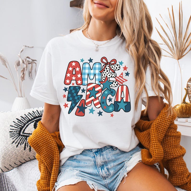 May include: White t-shirt featuring a colourful "AMERICA" design. The graphic includes stars, stripes, and a lightning bolt in red, white, and blue. A casual, everyday wear item.