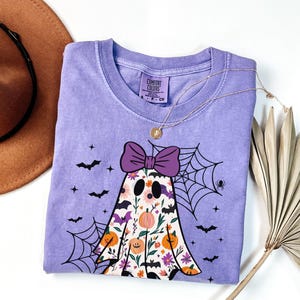 May include: A lavender Comfort Colors t-shirt with a ghost design, purple bow, floral details, and bats. A gold necklace and a brown hat are also visible. The shirt has a crew neck and short sleeves, ideal for Halloween.