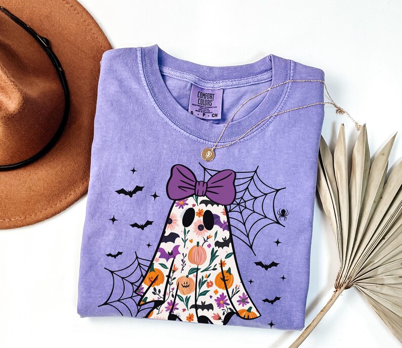 May include: A lavender Comfort Colors t-shirt with a ghost design, purple bow, floral details, and bats. A gold necklace and a brown hat are also visible. The shirt has a crew neck and short sleeves, ideal for Halloween.
