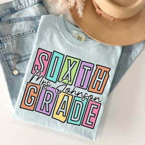 May include: A light blue t-shirt with the words "SIXTH Mrs. Johnson GRADE" in a colorful, block letter design. The shirt is laid on top of a pair of light blue jeans and a tan hat.