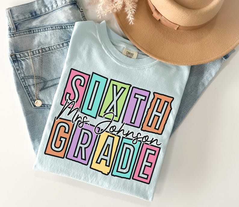 May include: A light blue t-shirt with the words "SIXTH Mrs. Johnson GRADE" in a colorful, block letter design. The shirt is laid on top of a pair of light blue jeans and a tan hat.