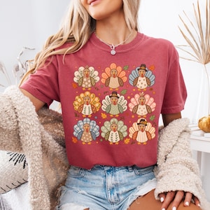 May include: A brick red crewneck t-shirt featuring a grid of nine illustrated turkeys. Each turkey has a unique patterned tail, including floral, plaid, and polka dot designs, with some wearing small hats, surrounded by scattered autumn leaves.
