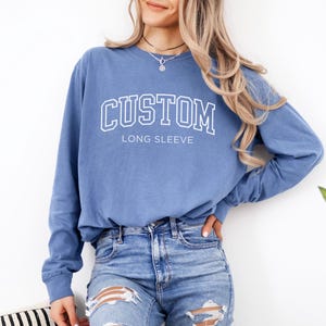 Op de afbeelding: Een blauw sweatshirt met lange mouwen met het woord "CUSTOM" in witte blokletters. Onder het woord "CUSTOM" staat de tekst "LONG SLEEVE" in witte blokletters. De sweatshirt heeft een effen blauwe kleur.