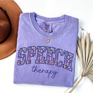 May include: A lilac Comfort Colors t-shirt with the word "SPEECH" in a floral design and the word "therapy" underneath. A gold necklace and a brown hat are next to the shirt. A dried palm leaf is in the background.