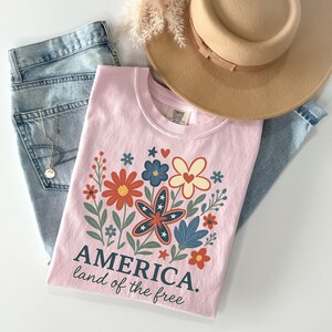 Peut inclure: T-shirt rose avec un motif floral compos&eacute; d'&eacute;toiles rouges, blanches et bleues. Le texte "AMERICA. land of the free" est imprim&eacute; sous le motif.