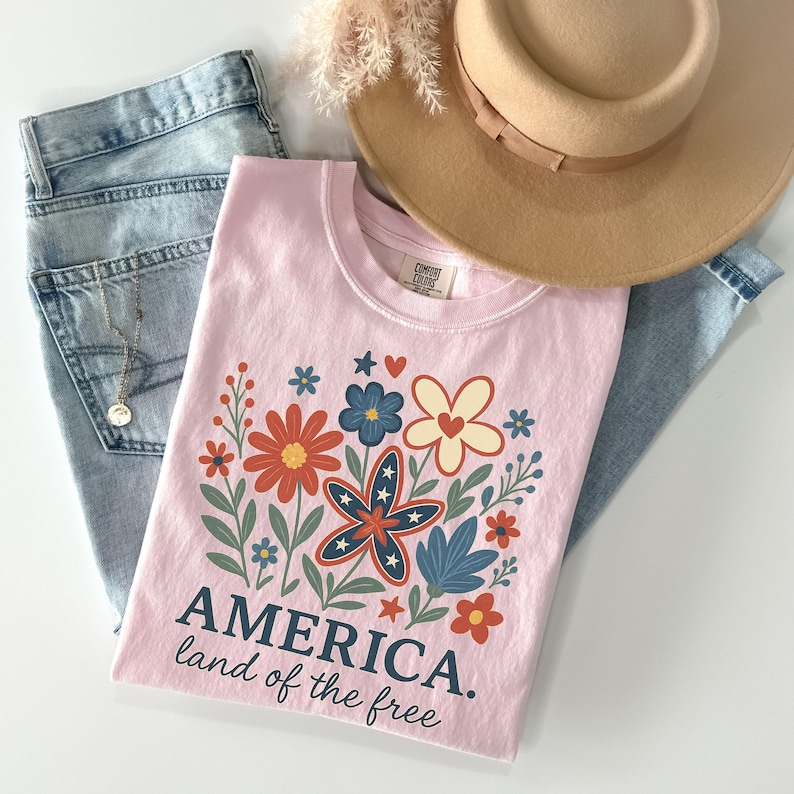 Peut inclure: T-shirt rose avec un motif floral compos&eacute; d'&eacute;toiles rouges, blanches et bleues. Le texte "AMERICA. land of the free" est imprim&eacute; sous le motif.