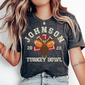 Op de afbeelding: Een donkergrijs T-shirt met een vintage-stijl afbeelding van een kalkoen die voetballen als veren draagt. De tekst "JOHNSON 20 23 TURKEY BOWL" is op het shirt gedrukt.