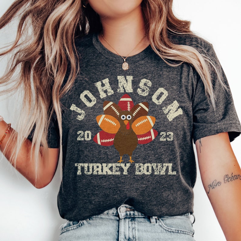 Op de afbeelding: Een donkergrijs T-shirt met een vintage-stijl afbeelding van een kalkoen die voetballen als veren draagt. De tekst "JOHNSON 20 23 TURKEY BOWL" is op het shirt gedrukt.