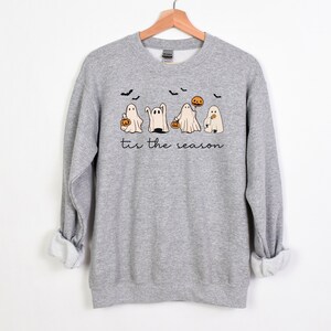 May include: A heather gray sweatshirt with a Halloween-themed graphic. The design features five cartoon ghosts, bats, and a pumpkin, with the phrase "tis the season" in a script font. The sweatshirt is displayed on a wooden hanger.