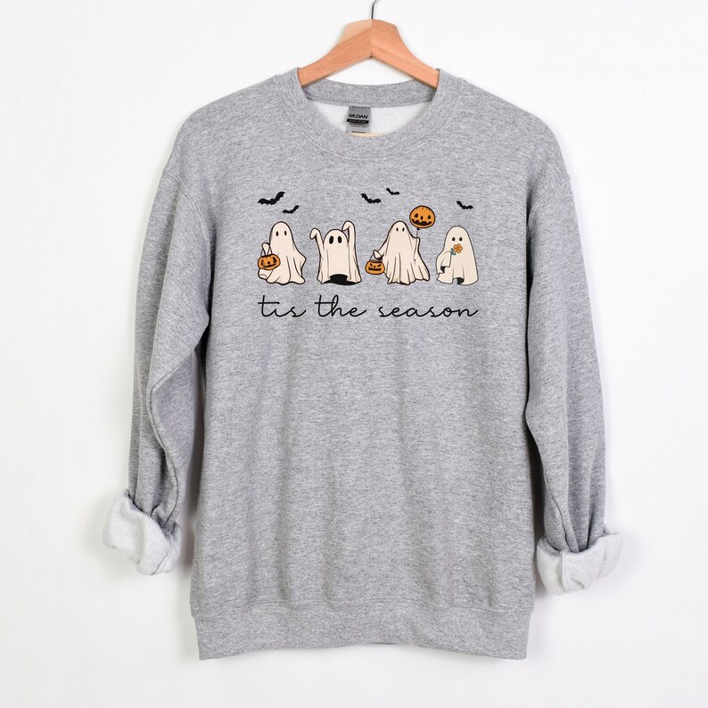 May include: A heather gray sweatshirt with a Halloween-themed graphic. The design features five cartoon ghosts, bats, and a pumpkin, with the phrase "tis the season" in a script font. The sweatshirt is displayed on a wooden hanger.