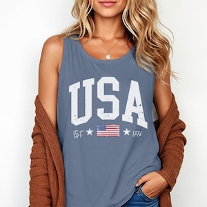 May include: A blue sleeveless tank top with a white distressed graphic that says "USA" with a small American flag and the text "Est. 1776".