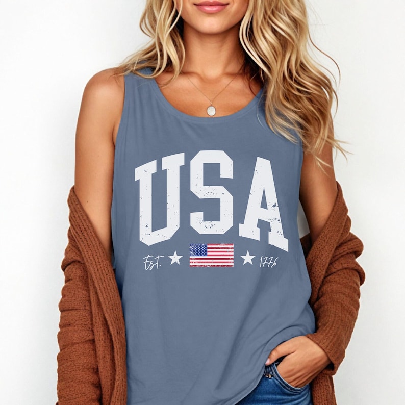 May include: A blue sleeveless tank top with a white distressed graphic that says "USA" with a small American flag and the text "Est. 1776".