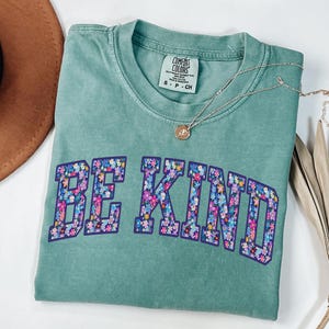 May include: A seafoam green Comfort Colors t-shirt with the words "BE KIND" in a floral design. A silver necklace with a small pendant rests on the shirt. A brown hat and dried palm leaf are in the background.