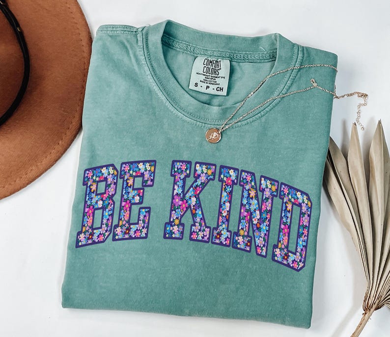 May include: A seafoam green Comfort Colors t-shirt with the words "BE KIND" in a floral design. A silver necklace with a small pendant rests on the shirt. A brown hat and dried palm leaf are in the background.