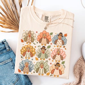 May include: A cream-colored t-shirt featuring a graphic print of nine stylized turkeys arranged in a grid. Each turkey has a unique patterned tail, including floral, plaid, and heart designs. Some turkeys wear small hats, and small autumn leaves are scattered.