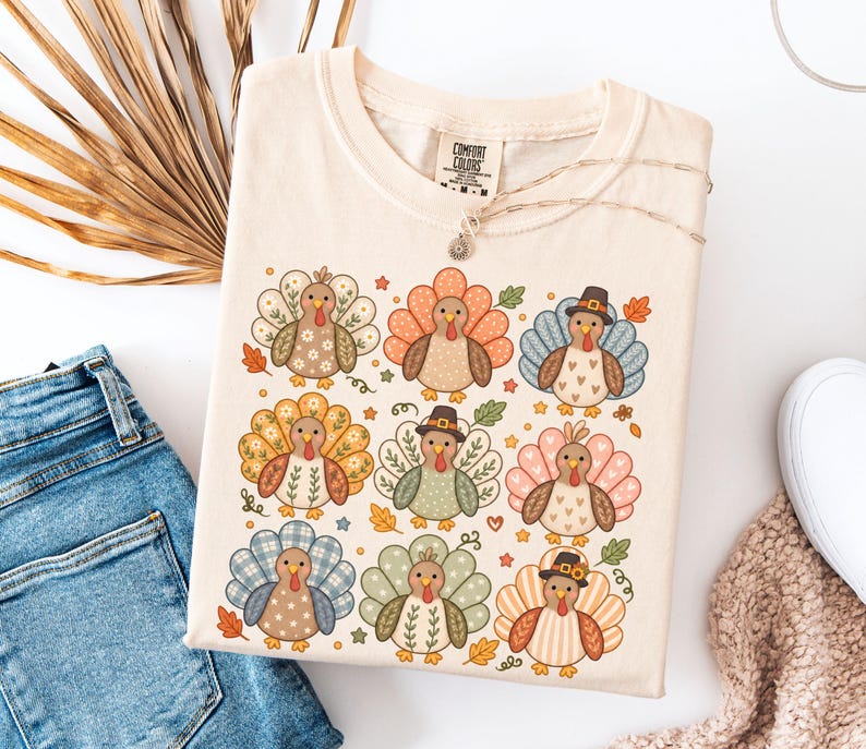 May include: A cream-colored t-shirt featuring a graphic print of nine stylized turkeys arranged in a grid. Each turkey has a unique patterned tail, including floral, plaid, and heart designs. Some turkeys wear small hats, and small autumn leaves are scattered.