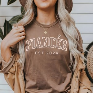 Puede incluir: Una camiseta marr&oacute;n con el texto "FIANC&Eacute;E EST 2024" en blanco.