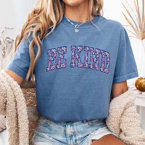 May include: A blue short-sleeved t-shirt with the words "BE KIND" in a floral design across the chest. The floral design includes pink, purple, and blue flowers. The shirt is made of a soft material.