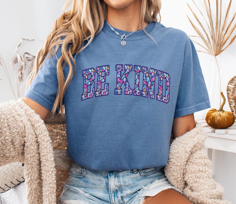 May include: A blue short-sleeved t-shirt with the words "BE KIND" in a floral design across the chest. The floral design includes pink, purple, and blue flowers. The shirt is made of a soft material.