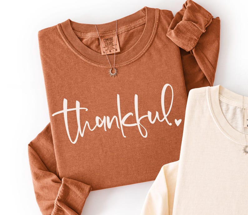 May include: A rust-colored long-sleeve shirt with the word "thankful" in white script. A silver necklace with a sunburst pendant is on the shirt. A cream-colored long-sleeve shirt is partially visible.