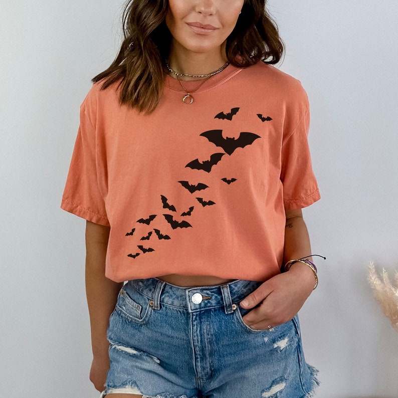 Puede incluir: Una camiseta de color naranja coral con una silueta negra de murci&eacute;lagos volando en diagonal por la parte delantera de la camiseta.
