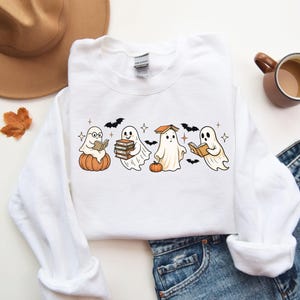 Puede incluir: Sudadera blanca de cuello redondo con un dise&ntilde;o de Halloween. El dise&ntilde;o presenta cuatro fantasmas de dibujos animados leyendo libros, con calabazas y murci&eacute;lagos. Un estilo c&oacute;modo e informal.