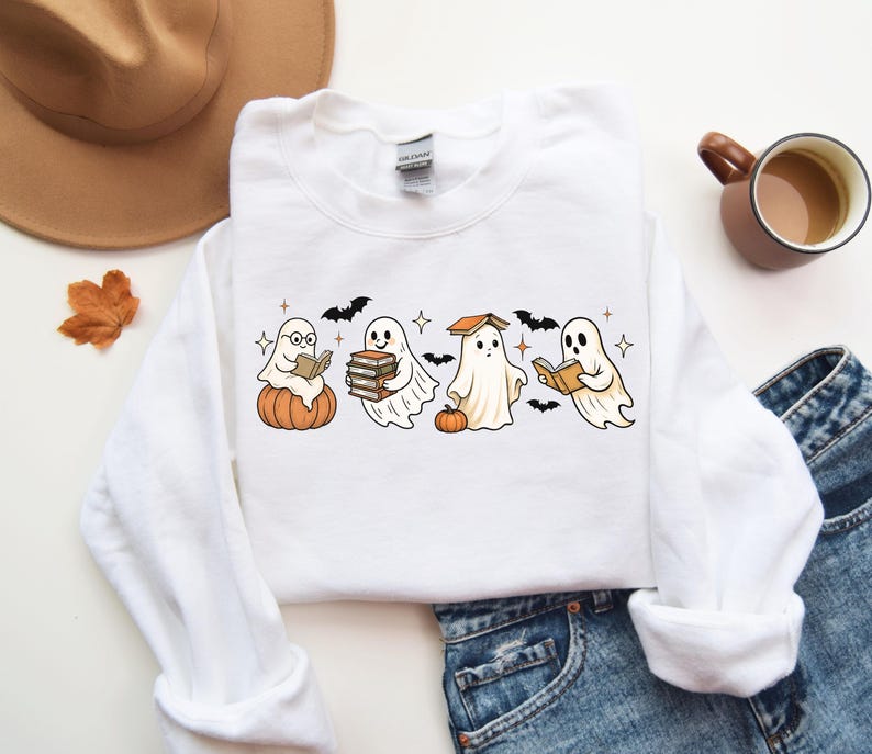 Puede incluir: Sudadera blanca de cuello redondo con un dise&ntilde;o de Halloween. El dise&ntilde;o presenta cuatro fantasmas de dibujos animados leyendo libros, con calabazas y murci&eacute;lagos. Un estilo c&oacute;modo e informal.