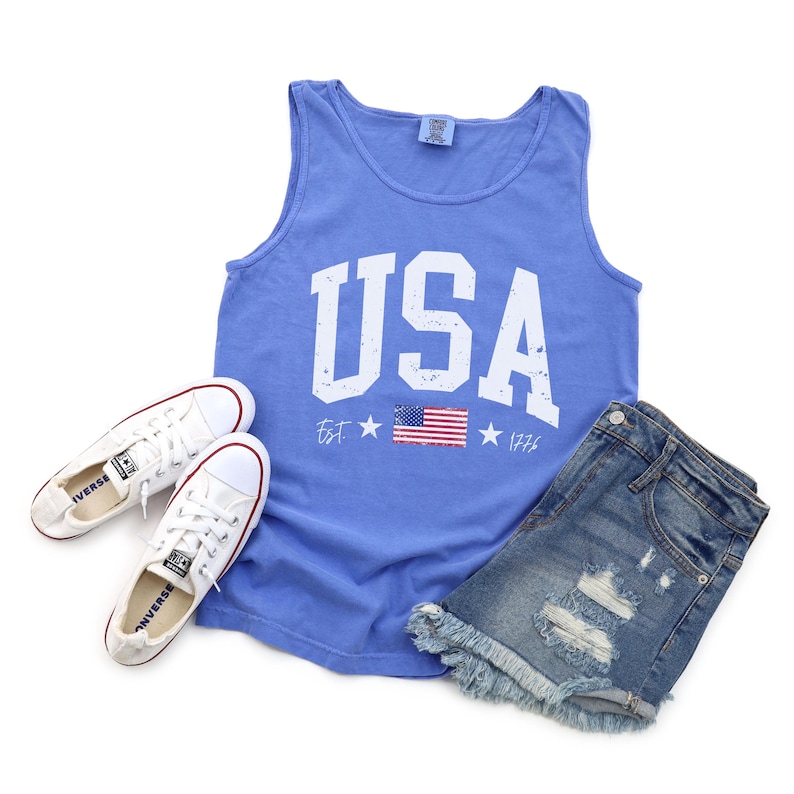 May include: A blue tank top with a white distressed "USA" graphic and a small American flag. The text reads "Est. 1776".