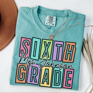 May include: A turquoise Comfort Colors t-shirt with the words "SIXTH Mrs. Johnson GRADE" in a colorful, block letter design. A gold necklace with a pendant is draped across the shirt. The shirt is folded on a white surface.