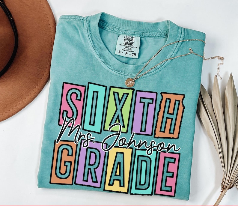 May include: A turquoise Comfort Colors t-shirt with the words "SIXTH Mrs. Johnson GRADE" in a colorful, block letter design. A gold necklace with a pendant is draped across the shirt. The shirt is folded on a white surface.