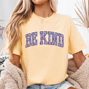 May include: A light yellow t-shirt with the words "BE KIND" in a floral design. The text is in a dark blue, arched font. The shirt has short sleeves and a crew neck.