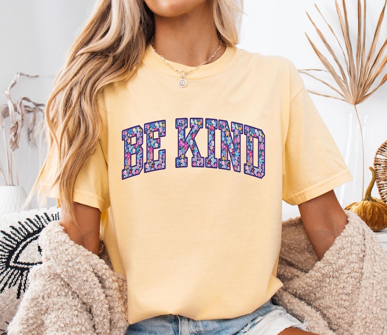 May include: A light yellow t-shirt with the words "BE KIND" in a floral design. The text is in a dark blue, arched font. The shirt has short sleeves and a crew neck.