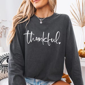 May include: Dark gray long-sleeve sweatshirt with the word "thankful" in white script. The word has a small heart at the end. The sweatshirt is a casual style, suitable for fall or winter wear.