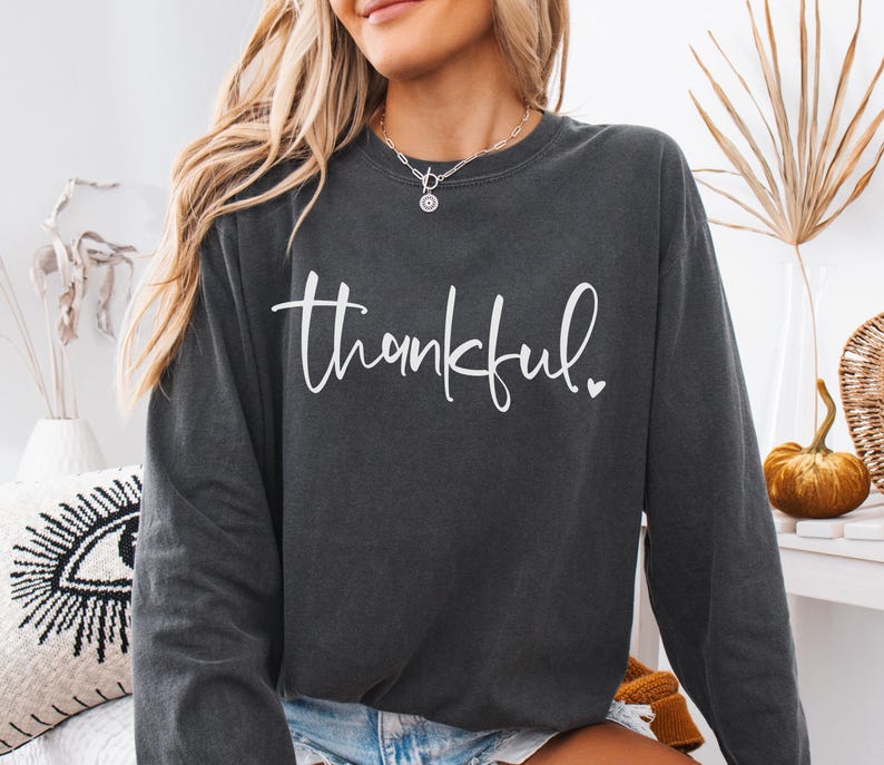 May include: Dark gray long-sleeve sweatshirt with the word "thankful" in white script. The word has a small heart at the end. The sweatshirt is a casual style, suitable for fall or winter wear.