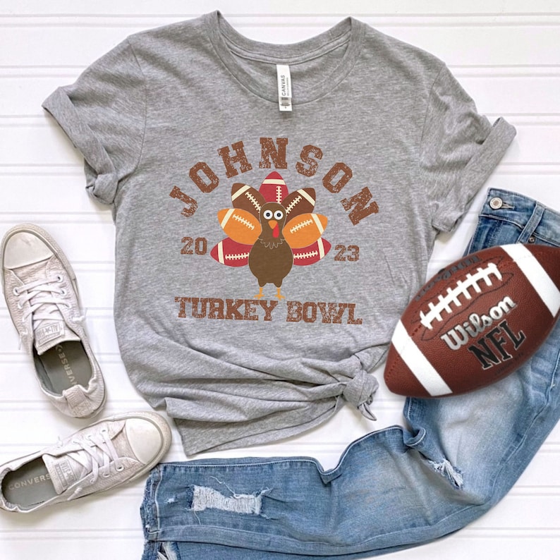 Puede incluir: Una camiseta gris con un dise&ntilde;o de pavo hecho de balones de f&uacute;tbol americano, el texto "JOHNSON 2023 TURKEY BOWL" en marr&oacute;n y un bal&oacute;n de f&uacute;tbol marr&oacute;n y blanco. La camiseta se combina con vaqueros azules y zapatillas blancas.
