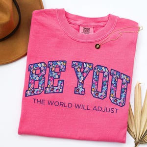 Pode incluir: Uma t-shirt de manga curta cor-de-rosa brilhante com o texto BE YOU num padr&atilde;o floral com contorno azul escuro, seguido pelo texto THE WORLD WILL ADJUST em letras azul escuro. A t-shirt tem uma etiqueta de pesco&ccedil;o da marca Comfort Colors.