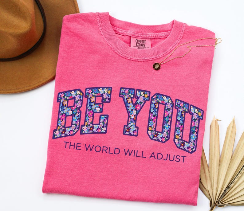 Pode incluir: Uma t-shirt de manga curta cor-de-rosa brilhante com o texto BE YOU num padr&atilde;o floral com contorno azul escuro, seguido pelo texto THE WORLD WILL ADJUST em letras azul escuro. A t-shirt tem uma etiqueta de pesco&ccedil;o da marca Comfort Colors.