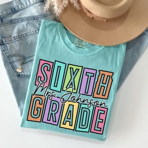 May include: A light blue t-shirt with the words "SIXTH GRADE Mrs. Johnson" in a colorful, block letter design. The shirt is paired with blue jeans and a tan hat, suggesting a casual, everyday style.