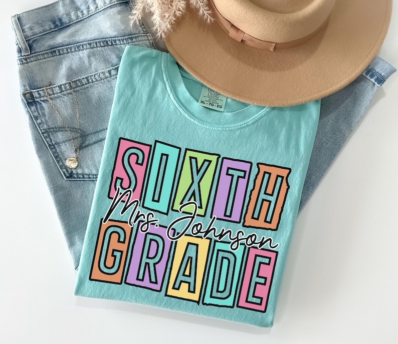 May include: A light blue t-shirt with the words "SIXTH GRADE Mrs. Johnson" in a colorful, block letter design. The shirt is paired with blue jeans and a tan hat, suggesting a casual, everyday style.